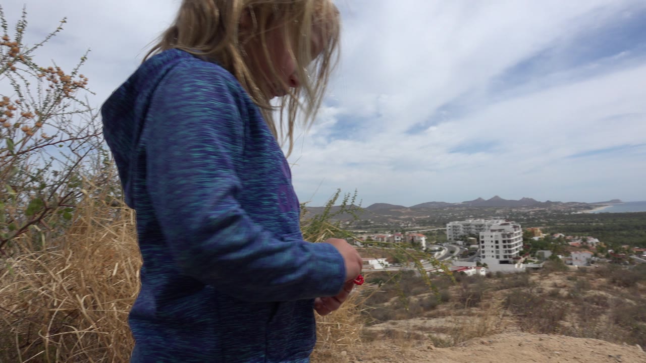 Blonde child in a blue hoodie stands close to the camera on a dry Los Cabos hillside, with scrub plants, town buildings, coastline, and mountains in the distance.