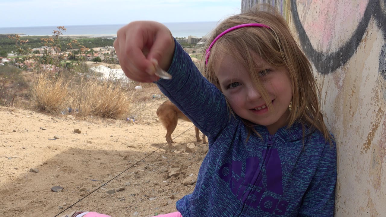 Young girl holds a small object toward the camera beside a painted wall.