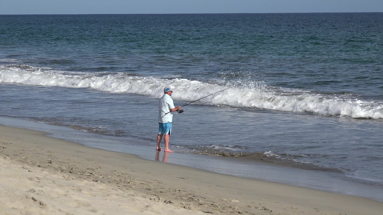 A man stands barefoot on wet sand in Los Cabos, Mexico, holding a fishing rod as a white wave breaks behind him near the start of the clip.