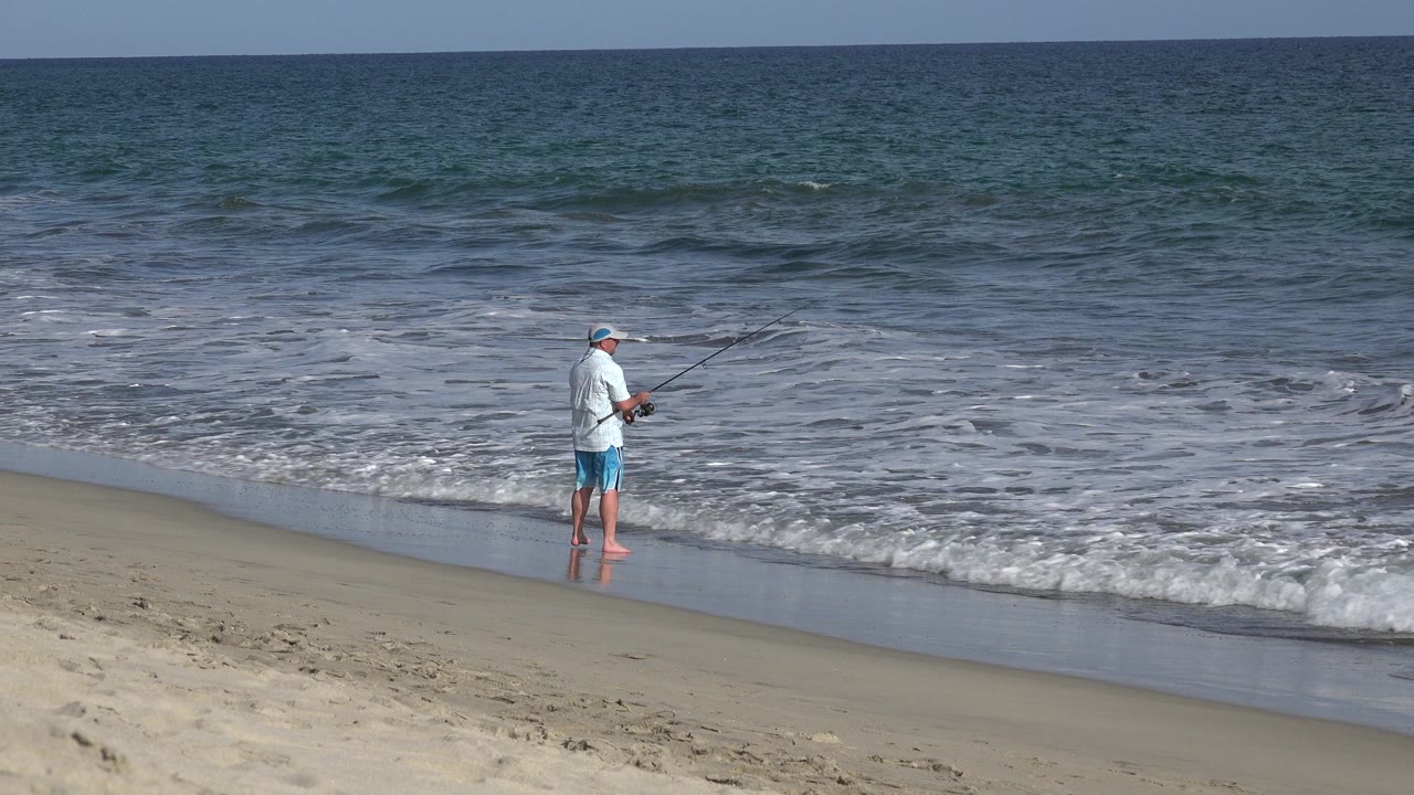 A lone man in a blue hat stands near the foamy shoreline in Los Cabos, Mexico, holding a fishing rod as the ocean fills the background.