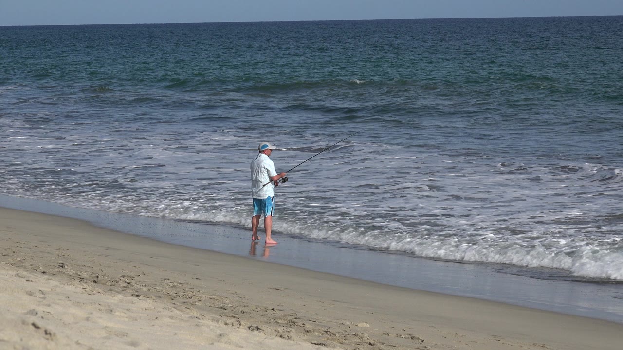 The man remains near the wet sand in Los Cabos, Mexico, with his fishing rod angled over the surf and a low wave breaking behind him.