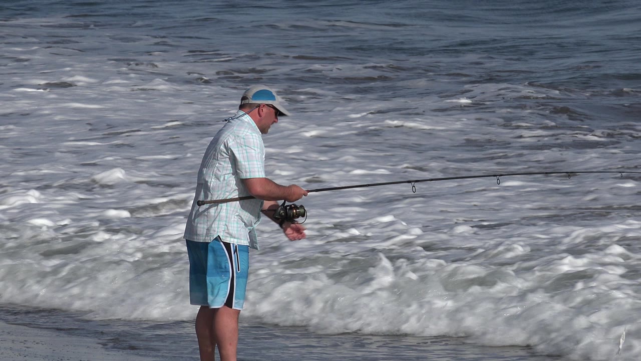 The fisherman remains near the foamy Los Cabos surf with the rod held low and stretched across the frame toward the ocean.