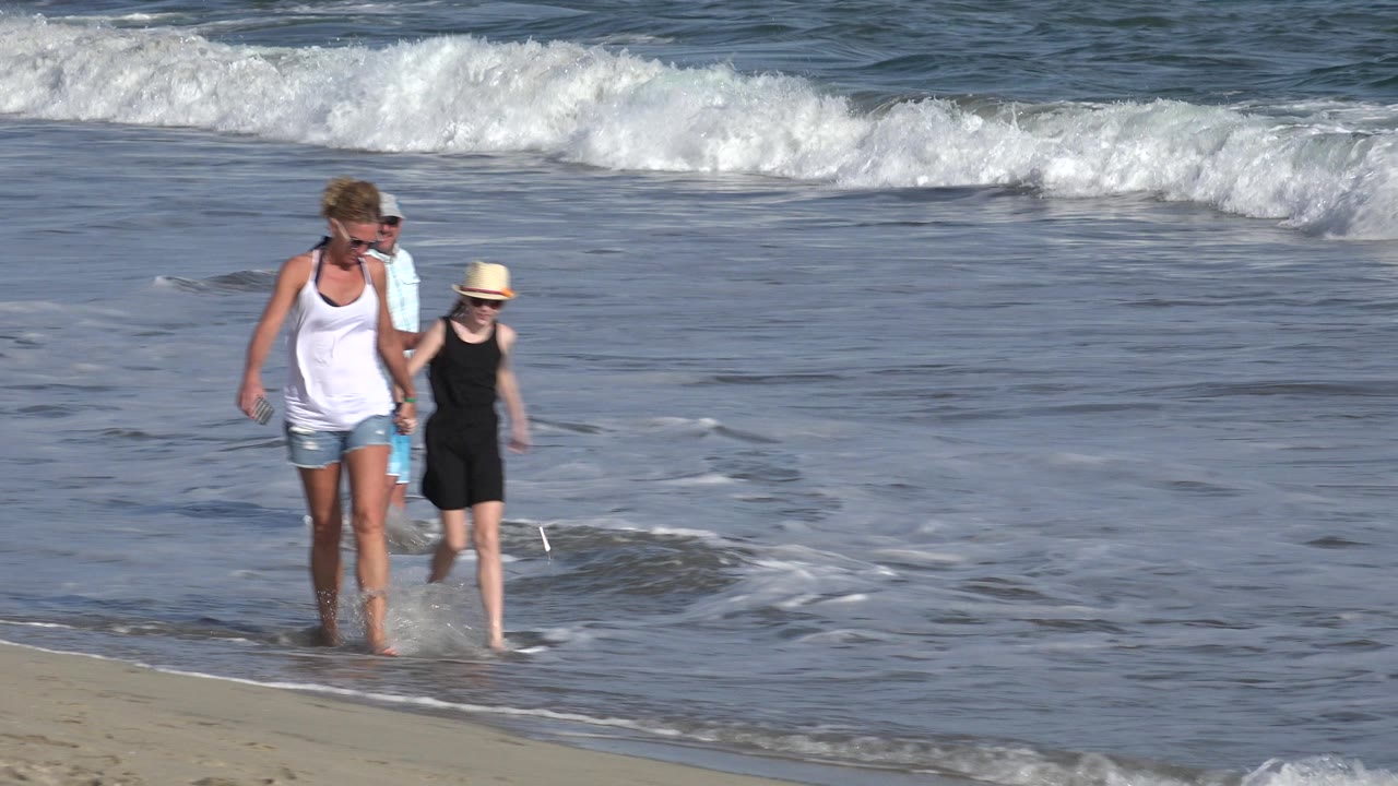 The woman and girl move forward through a thin wash of water on the Los Cabos beach, with white surf bands stretching across the background.