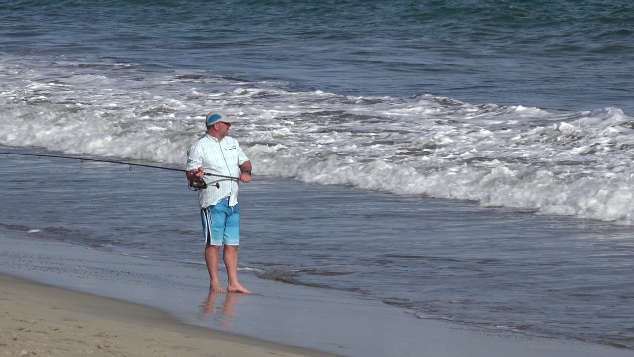 Man with fishing rod standing barefoot at the edge of the surf.