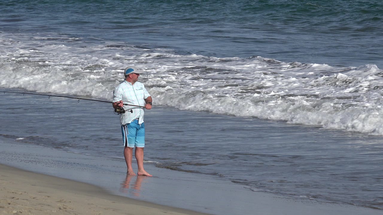 The man remains near the waterline in Los Cabos, Mexico, with the long rod angled across the frame and foamy surf spreading behind him.