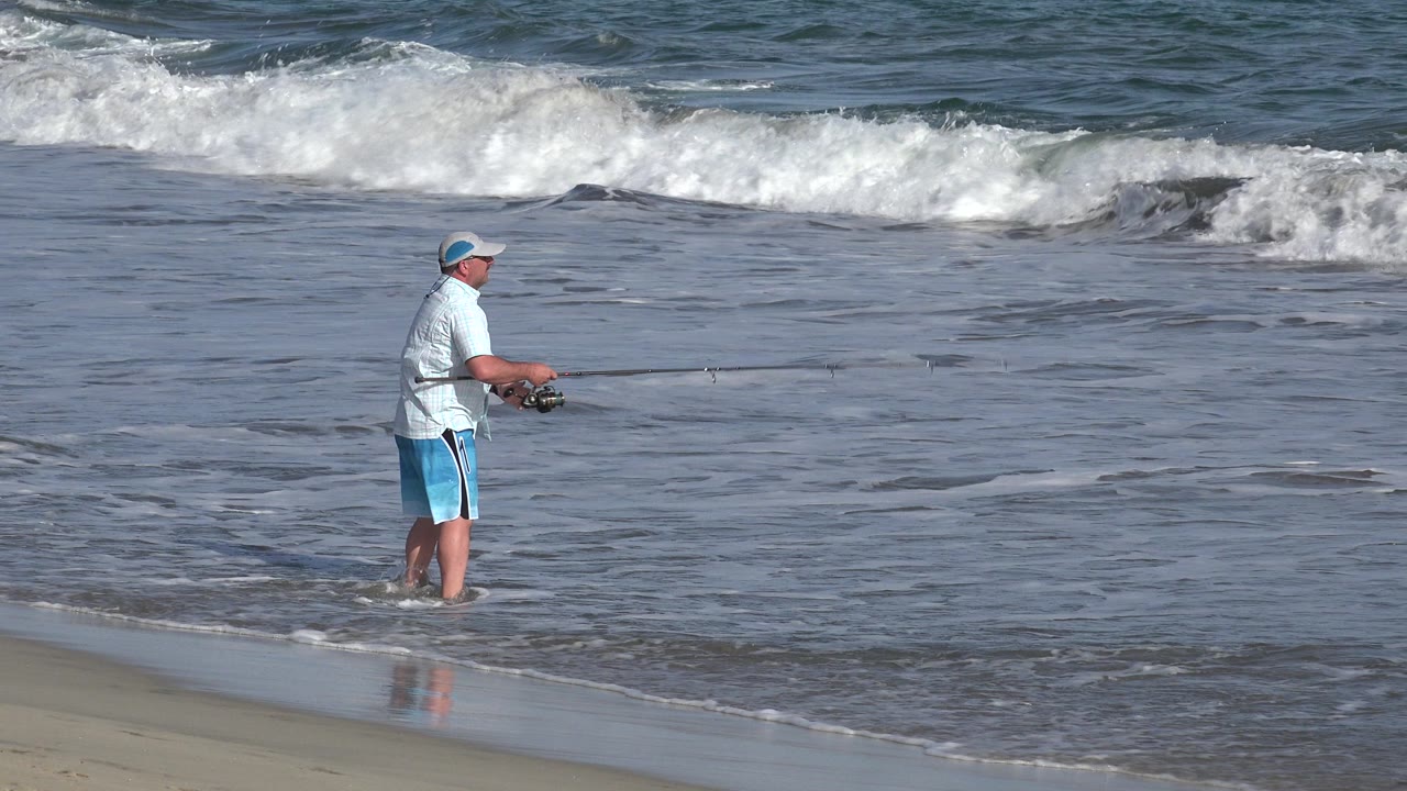The fisherman remains near the wet sand in Los Cabos, holding the rod forward while the foamy wave line spreads across the upper frame.