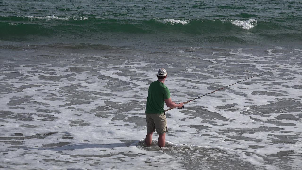 The fisherman remains turned toward the water, with his rod extended across white foam and darker green surf in Los Cabos, Mexico.