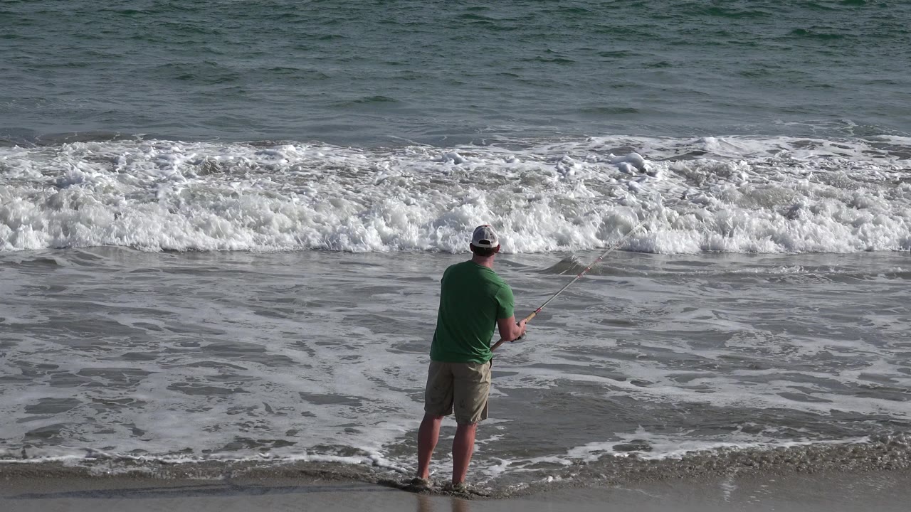 A man in a green shirt and shorts stands at the Los Cabos waterline with a fishing rod as a white wave breaks in front of him.