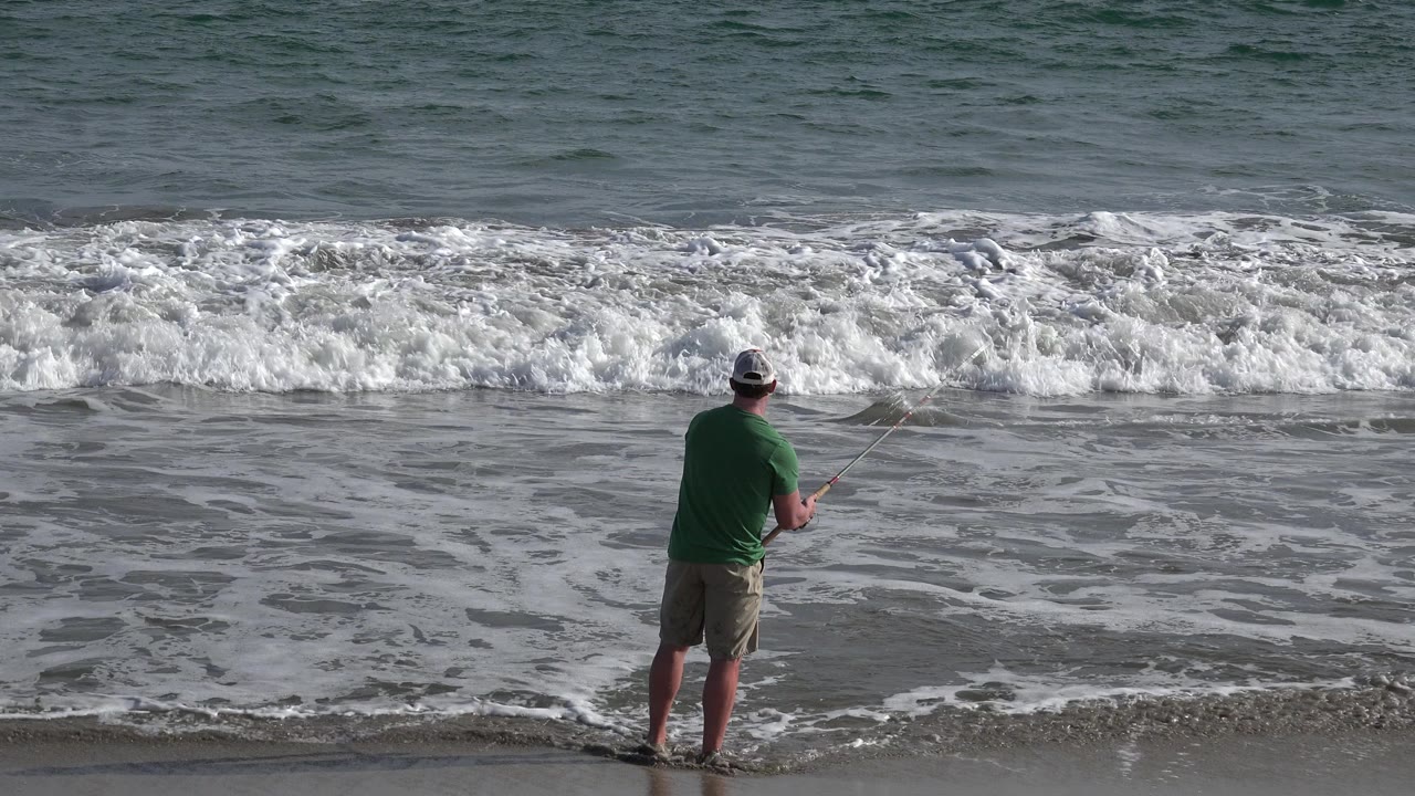 The fisherman remains near the Los Cabos shoreline while foam spreads across the wet sand and the rod points toward the incoming surf.