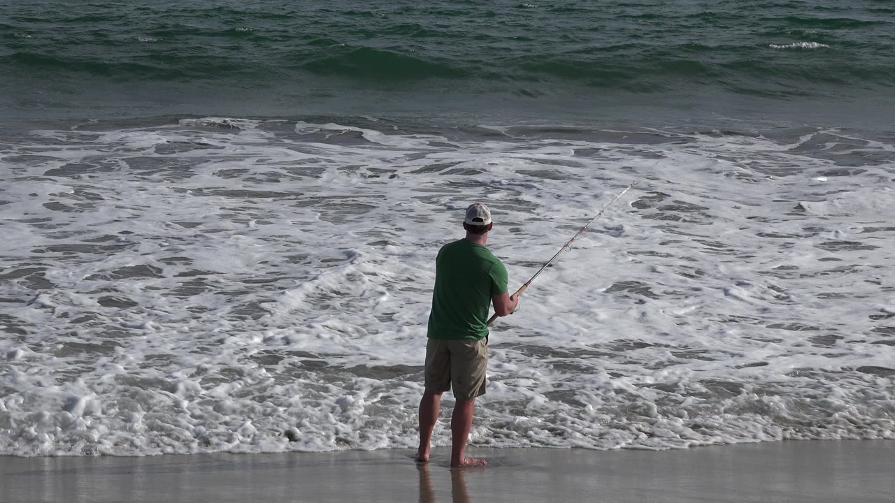 The fisherman remains centered on the wet beach in Los Cabos, holding his rod while white foam spreads across the shallow water in front of him.