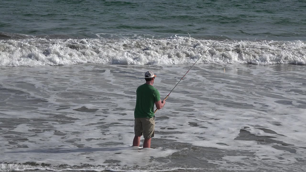 The fisherman remains ankle-deep in the surf in Los Cabos, Mexico, with his rod angled over a band of white water and darker ocean beyond.
