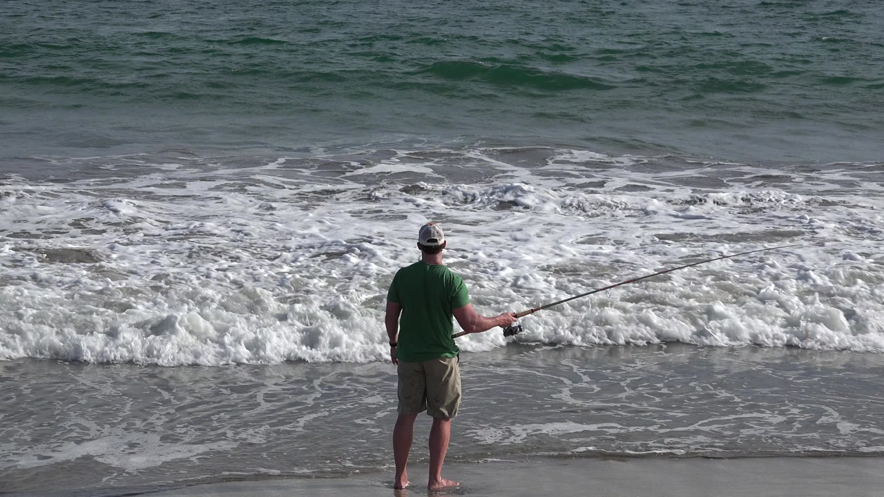 In Los Cabos, Mexico, a man in a green shirt and tan shorts stands near the waterline with a fishing rod as a foamy wave breaks in front of him.
