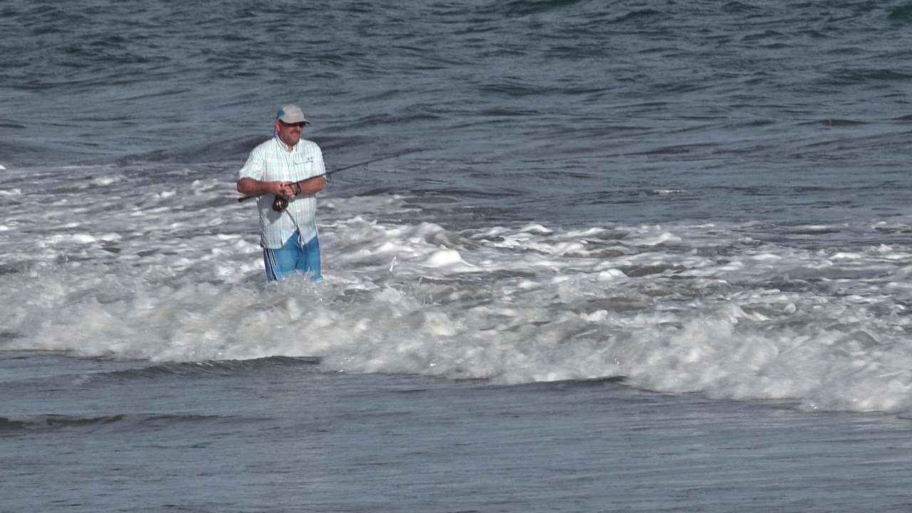 Adult fisherman in the Los Cabos surf holds his rod across his body while whitewater rolls across the lower frame.