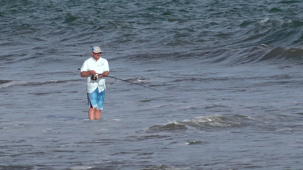 Man fishing in shallow surf with ocean waves behind him.