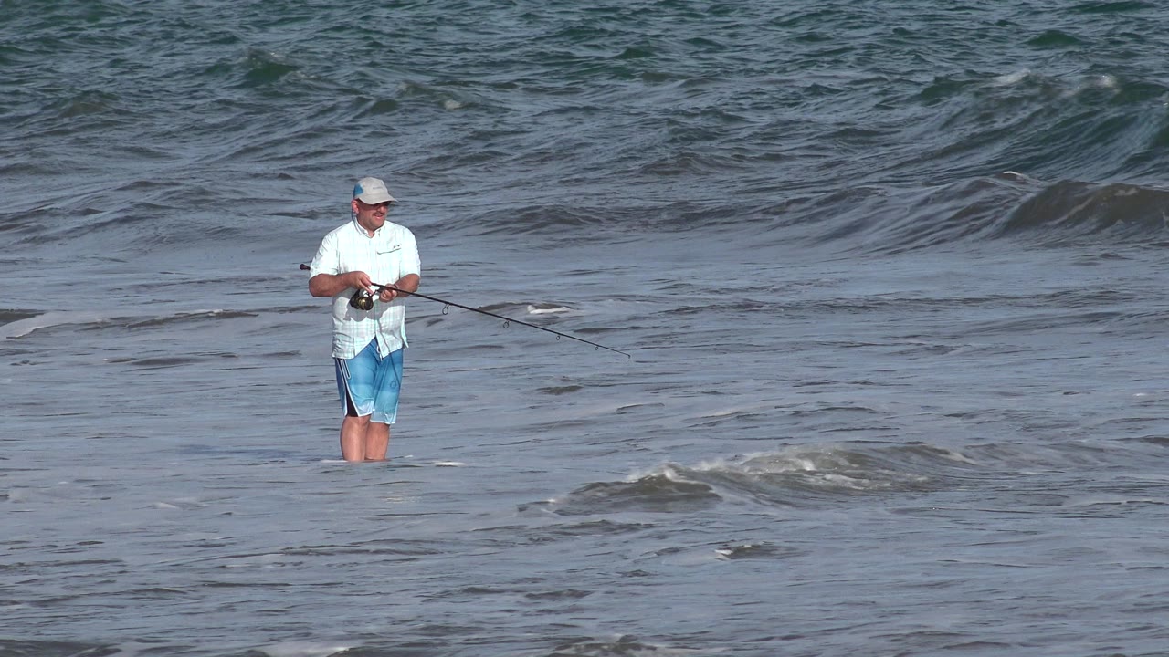 The fisherman remains centered in Los Cabos surf, holding his reel while low foam moves across the water in front of him.