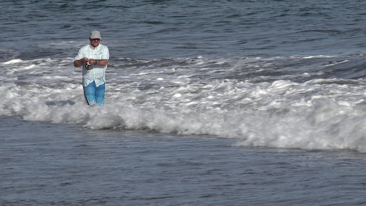 The man faces forward in shallow Los Cabos surf while a larger band of whitewater rolls between him and the camera.