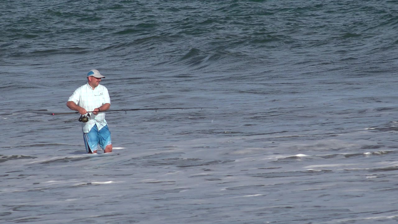 Man fishing knee-deep in surf with waves behind him.