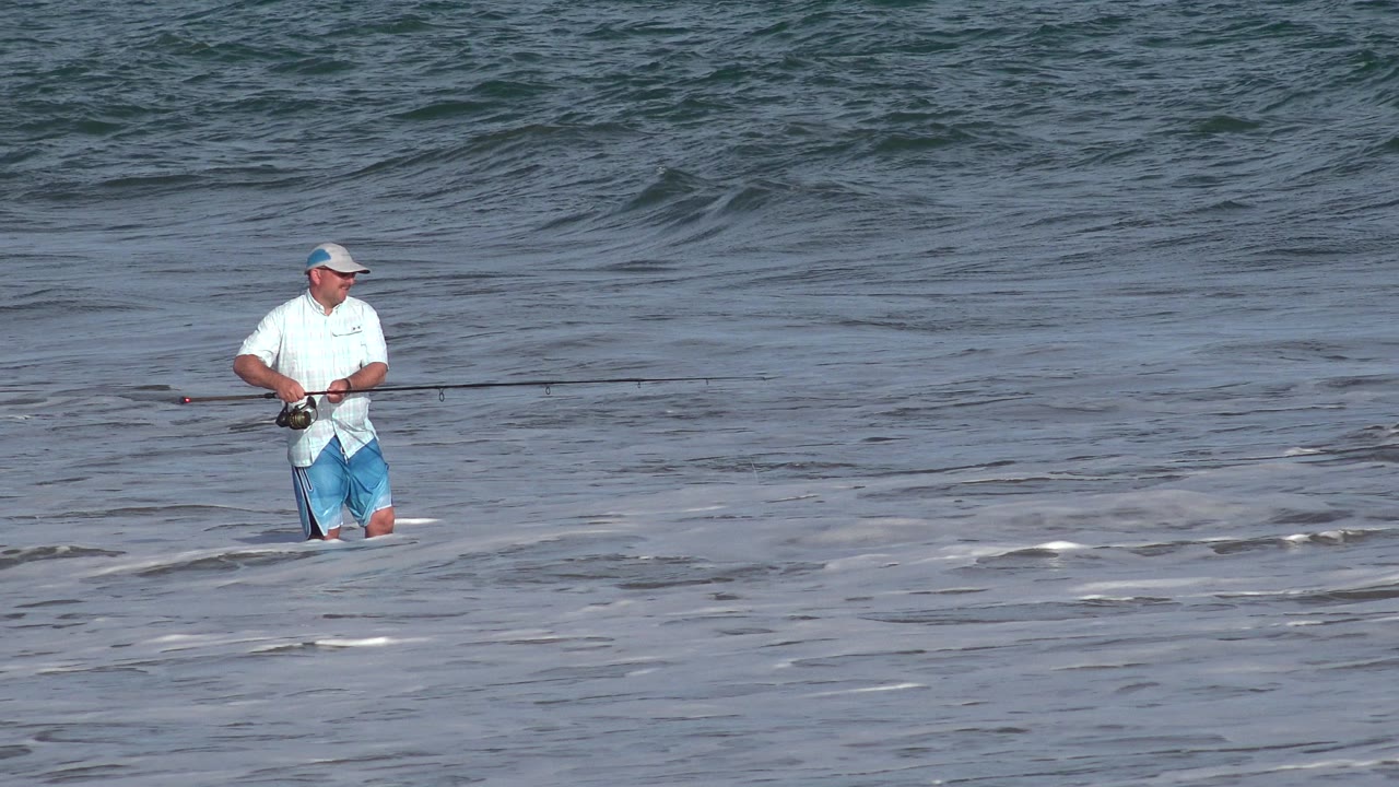 One second in, the fisherman remains knee-deep in Los Cabos surf, angled slightly to the side with his hands near the reel and a low wave line behind him.