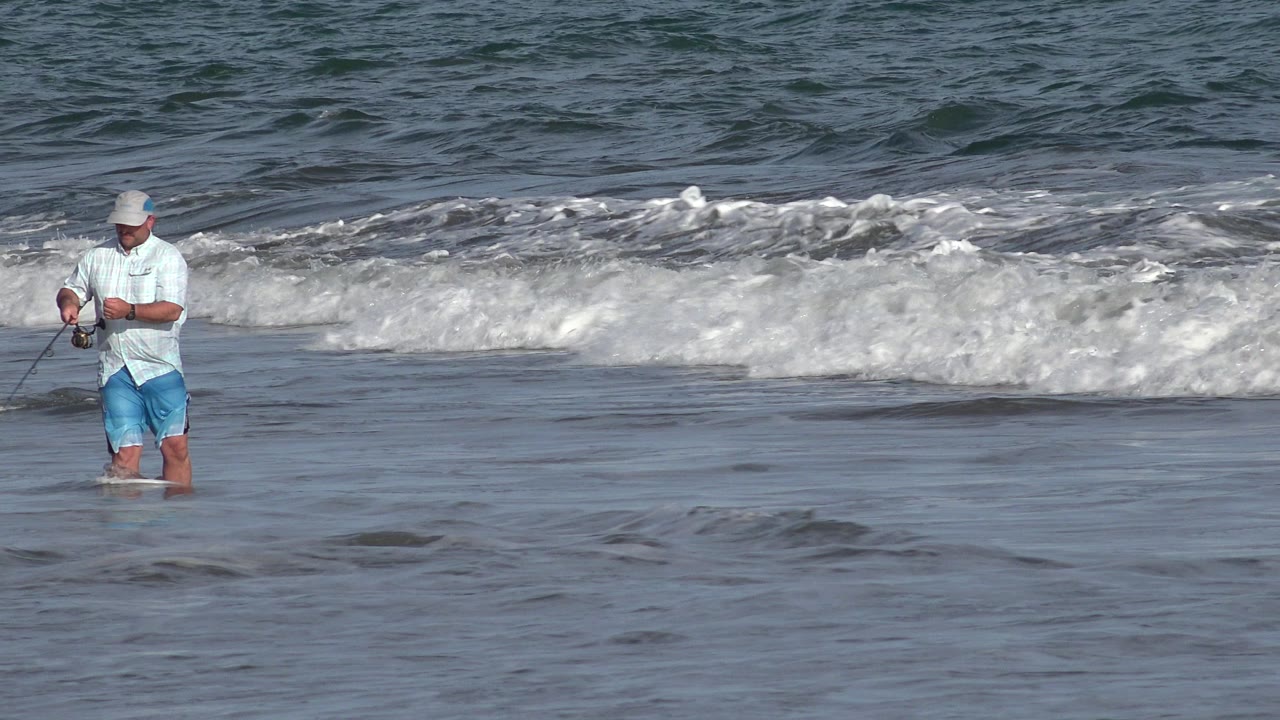The fisherman stands farther into the center of frame at a Los Cabos shoreline, holding his rod while foamy surf gathers behind him.