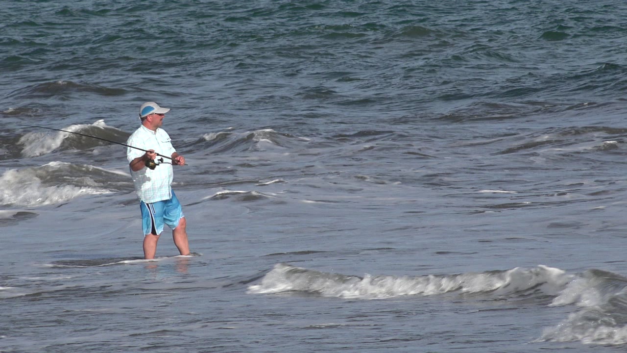 A man in a light shirt and blue shorts stands in shallow Los Cabos surf holding a fishing rod as a small wave breaks behind him.