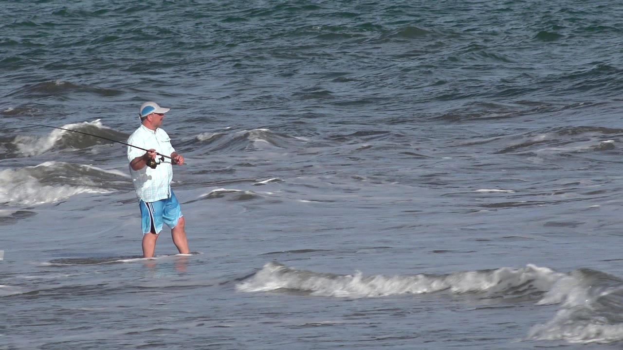 The fisherman remains knee-deep in Los Cabos water, facing across the surf while low gray-blue waves roll through the frame.