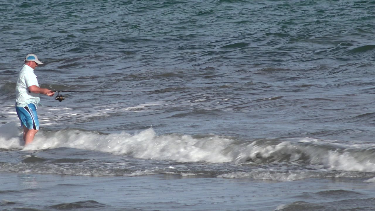 The adult angler stands farther into the frame with his rod angled over the gray-blue water as a band of white foam crosses the foreground in Los Cabos, Mexico.