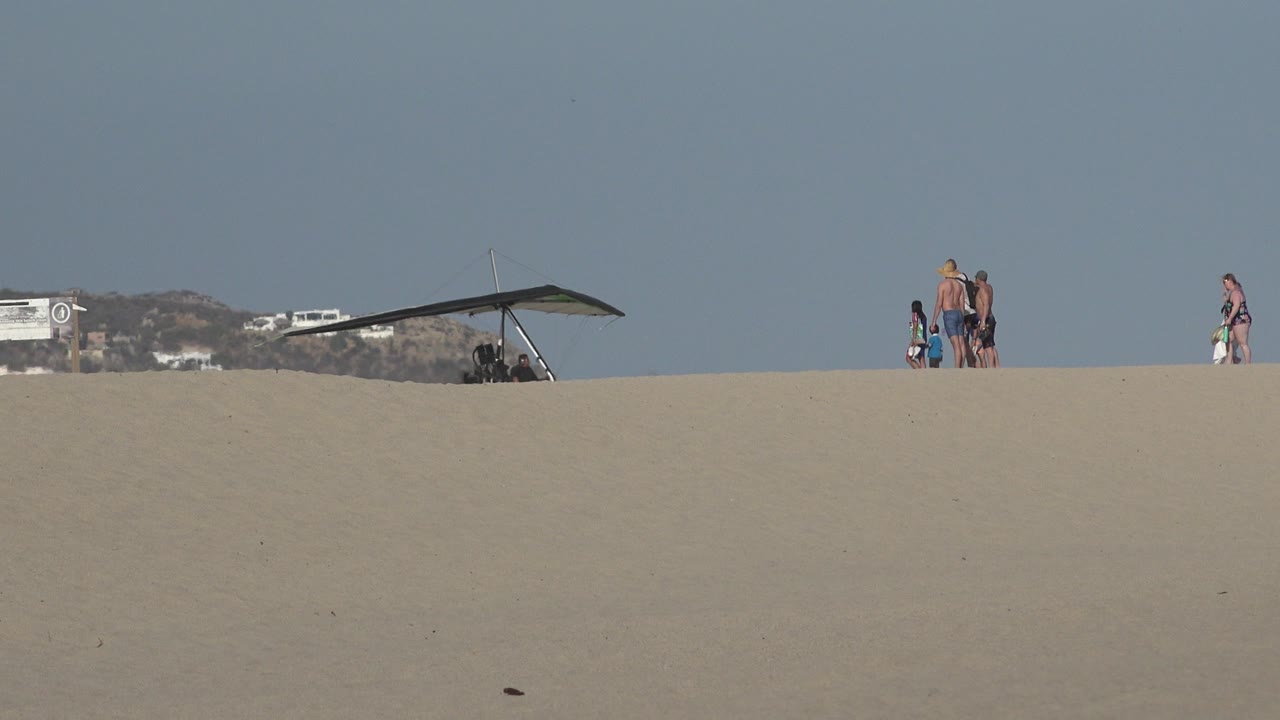 Los Cabos beach view with the hang glider canopy near the ridge, several vacationers walking or standing on the sand, and a pale blue sky dominating the frame.