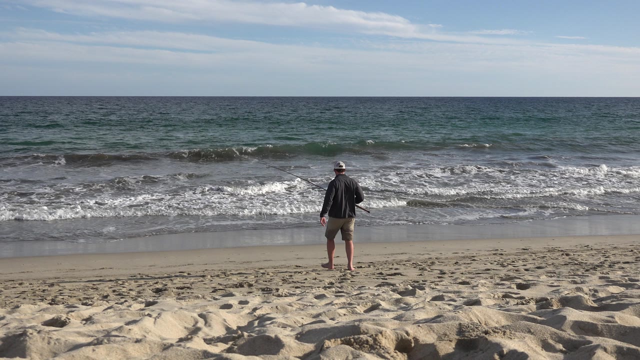 A man in a hat stands on a Los Cabos beach facing the blue-green ocean, with pale sand in the foreground and low surf breaking ahead.