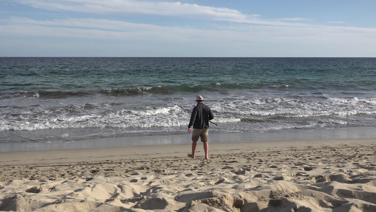 The man walks forward on the Los Cabos shoreline as small waves break across the middle of the frame and the horizon stays level in the distance.