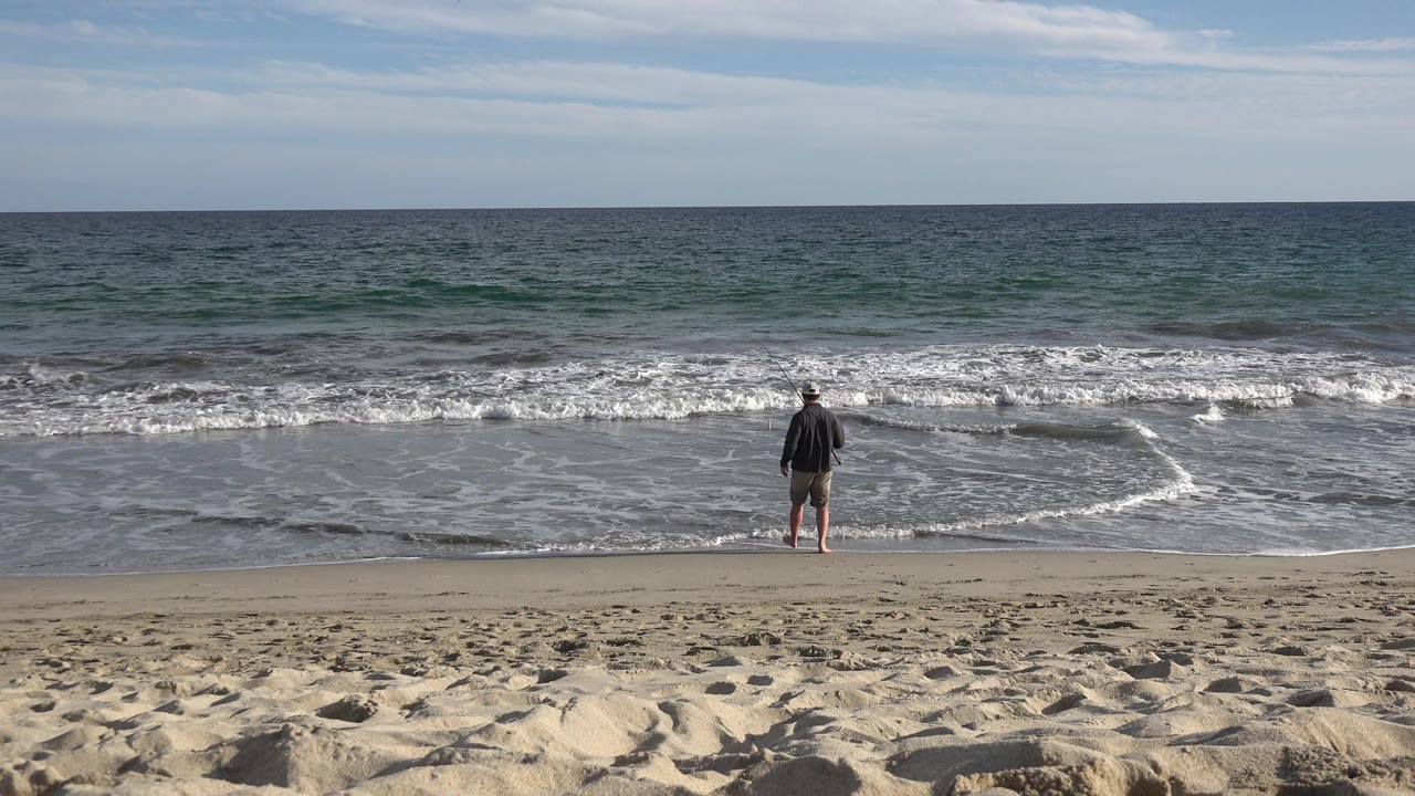 At 0:00, a man in a hat and shorts stands near the incoming surf with a fishing rod on a Los Cabos, Mexico beach, with pale sand in front and the ocean horizon behind him.