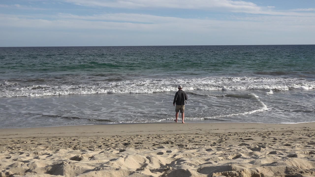 At 0:01, the fisherman remains centered near the waterline as a thin fishing line angles out across foamy waves on the Los Cabos shoreline.