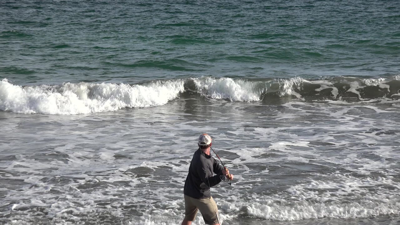 The fisherman remains low in the frame on the Los Cabos beach, holding his rod toward foamy water as another wave curls behind him.