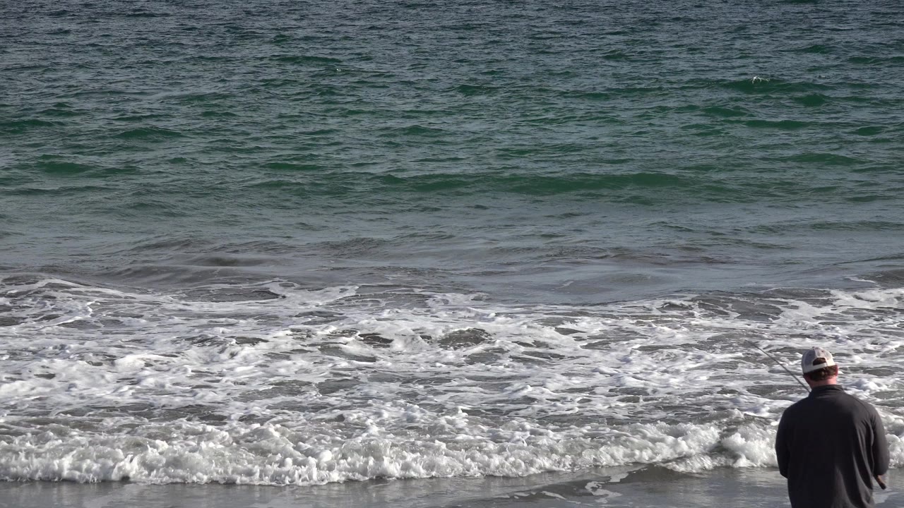 Man in a cap fishing at the shoreline with waves behind him.