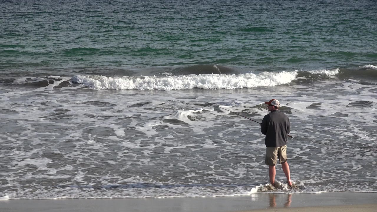 Man fishing at the edge of foamy surf.