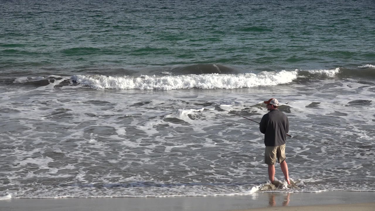 The fisherman remains facing the sea in Los Cabos, Mexico, with his rod angled left as a line of white surf breaks behind him.