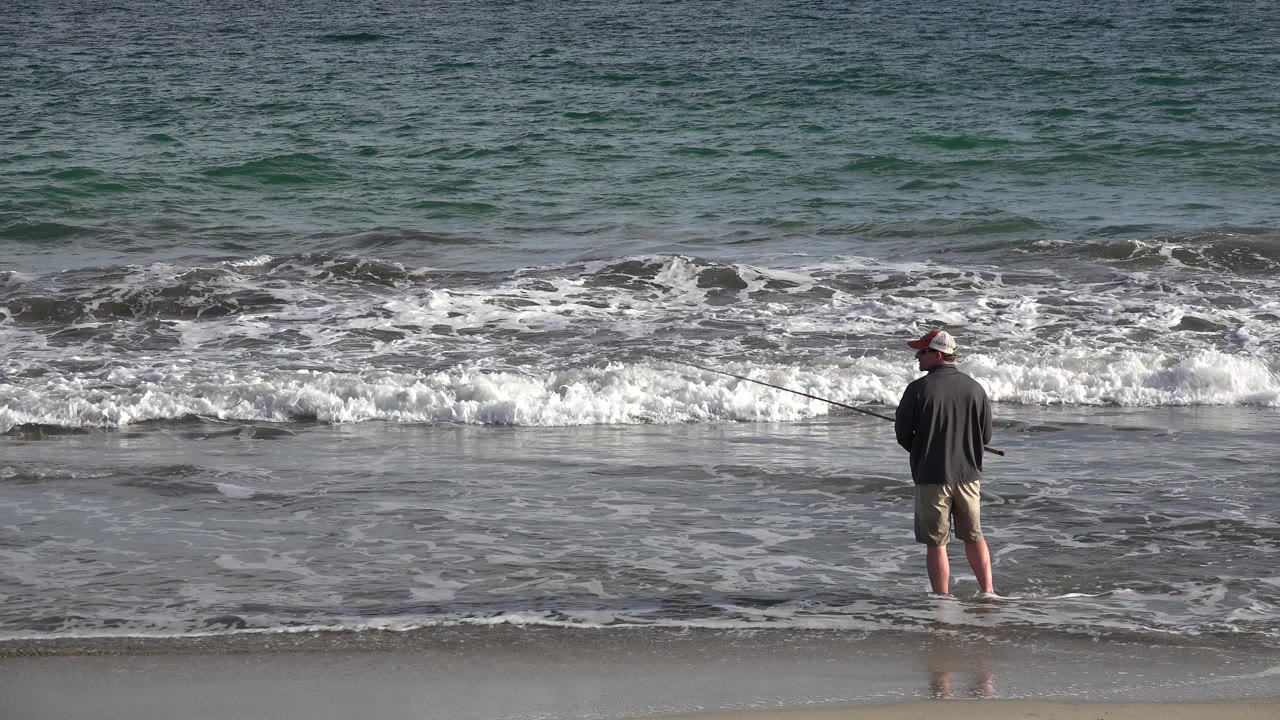 A man stands barefoot on wet sand in Los Cabos, Mexico, holding a fishing rod toward the surf as a foamy wave line breaks behind him.