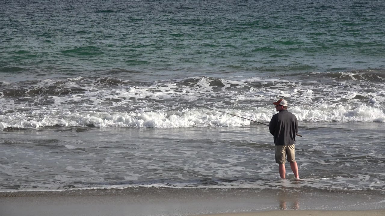 The fisherman remains centered on the wet beach in Los Cabos, Mexico, with his rod angled left and a brighter strip of broken surf behind him.