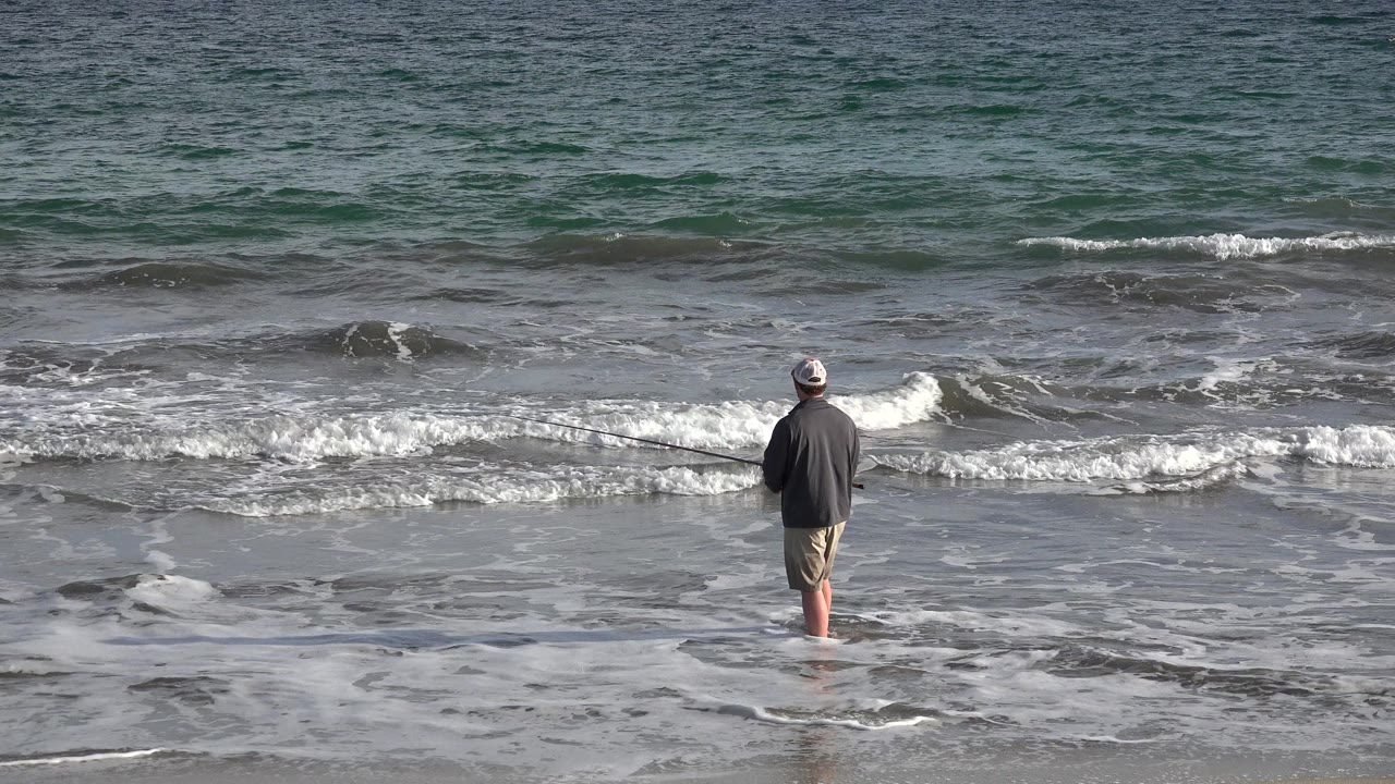 Man standing in shallow surf facing ocean waves.