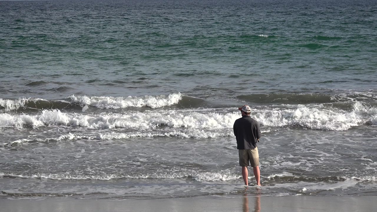 Man standing barefoot at the edge of breaking surf.