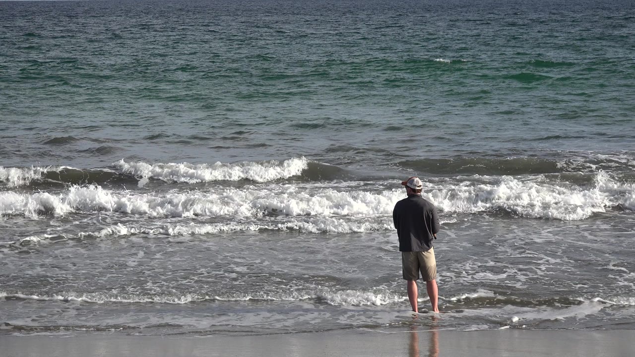 At 0:01, the man remains centered near the waterline in Los Cabos while a foamy wave spreads across the midground.