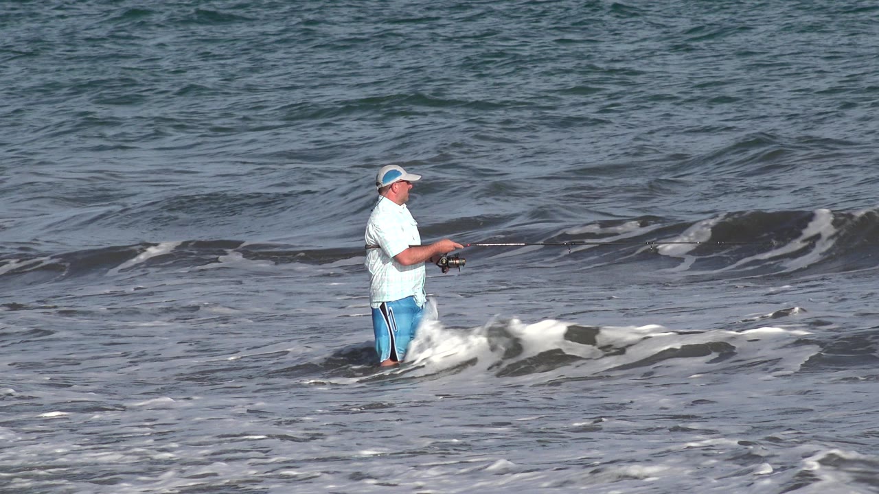 The fisherman remains near the center of the Los Cabos shoreline frame while white foam spreads around his legs and darker water sits behind him.