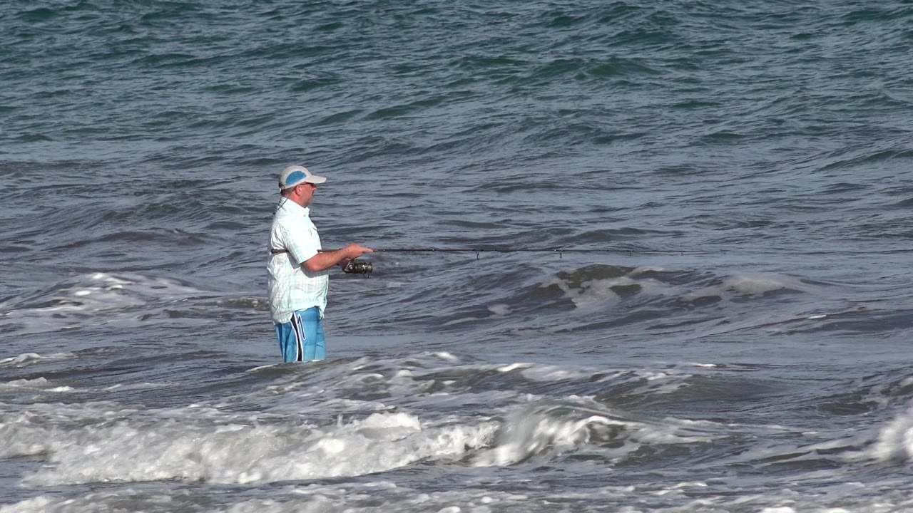 An adult man in a cap stands in shallow surf in Los Cabos, Mexico, holding a fishing rod as whitewater breaks across the foreground.