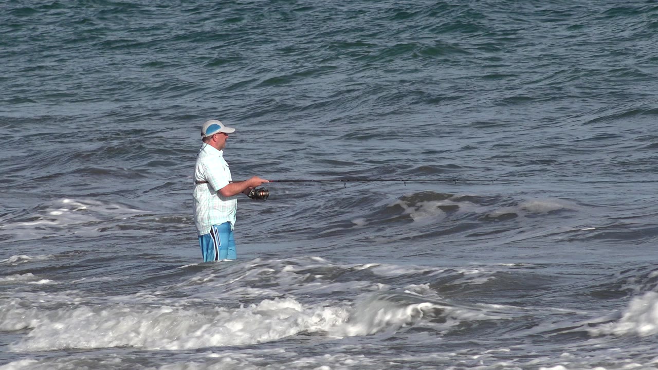 The fisherman remains in the shallow Los Cabos surf with his rod angled across the water and a foamy wave line rising in front of him.