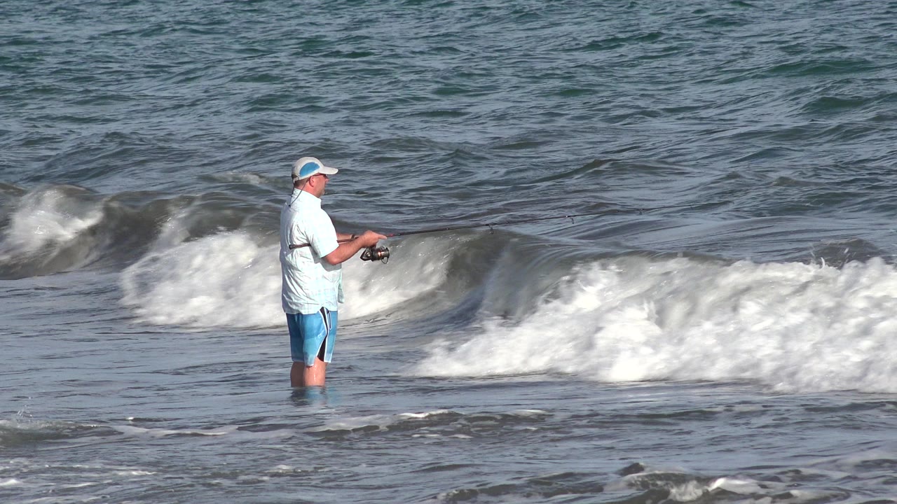 In Los Cabos, Mexico, a man in a pale shirt and blue shorts stands knee-deep in the surf with a fishing rod while a wave gathers behind him.