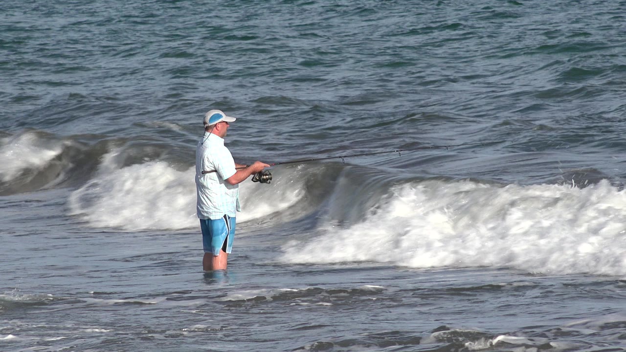 The Los Cabos shoreline frame holds on the fisherman as whitewater spreads across the wave line behind him and the rod angles out over the water.