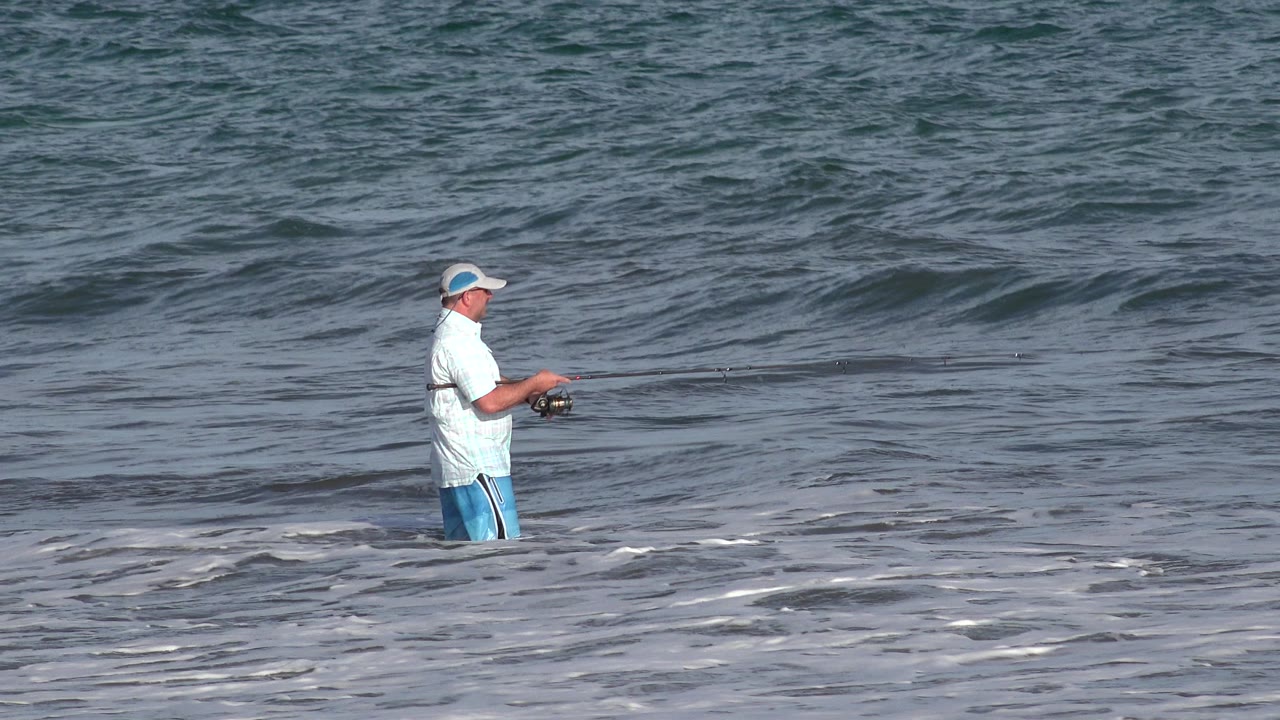 The fisherman remains knee-deep in the Los Cabos surf, turned sideways with his rod extended across the frame and darker water behind him.