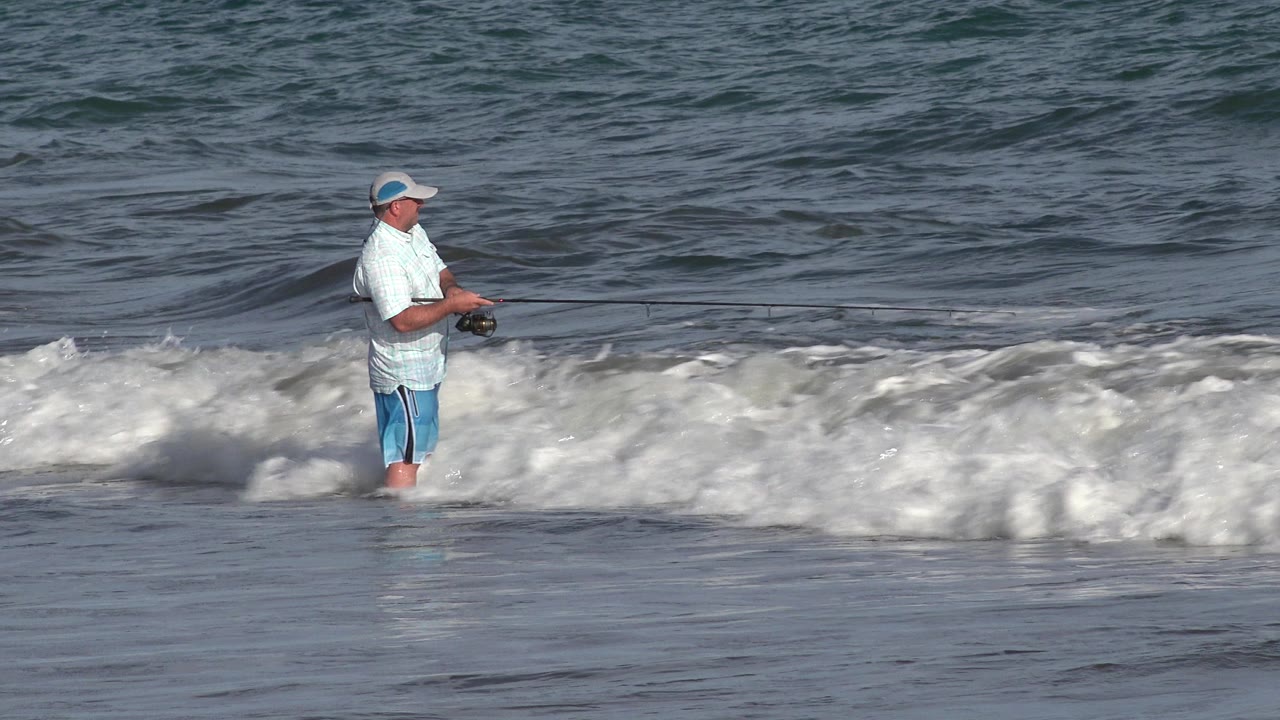 The fisherman remains in the shallow water in Los Cabos, Mexico, with a bright white wave crest spreading across the midground behind his legs.