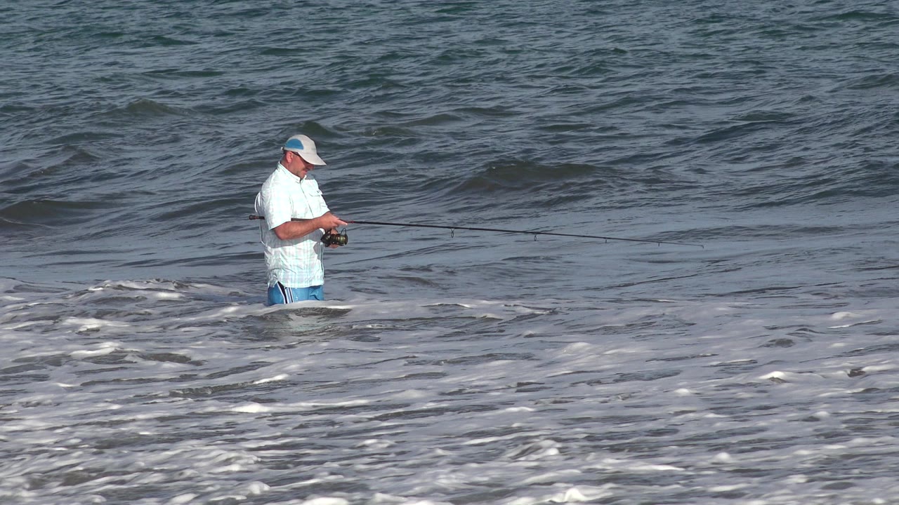 In Los Cabos, Mexico, the angler looks down toward his reel while standing in foamy water with darker ocean swell behind him.