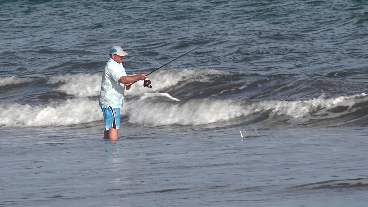 Man fishing in shallow surf as a small wave breaks behind him.