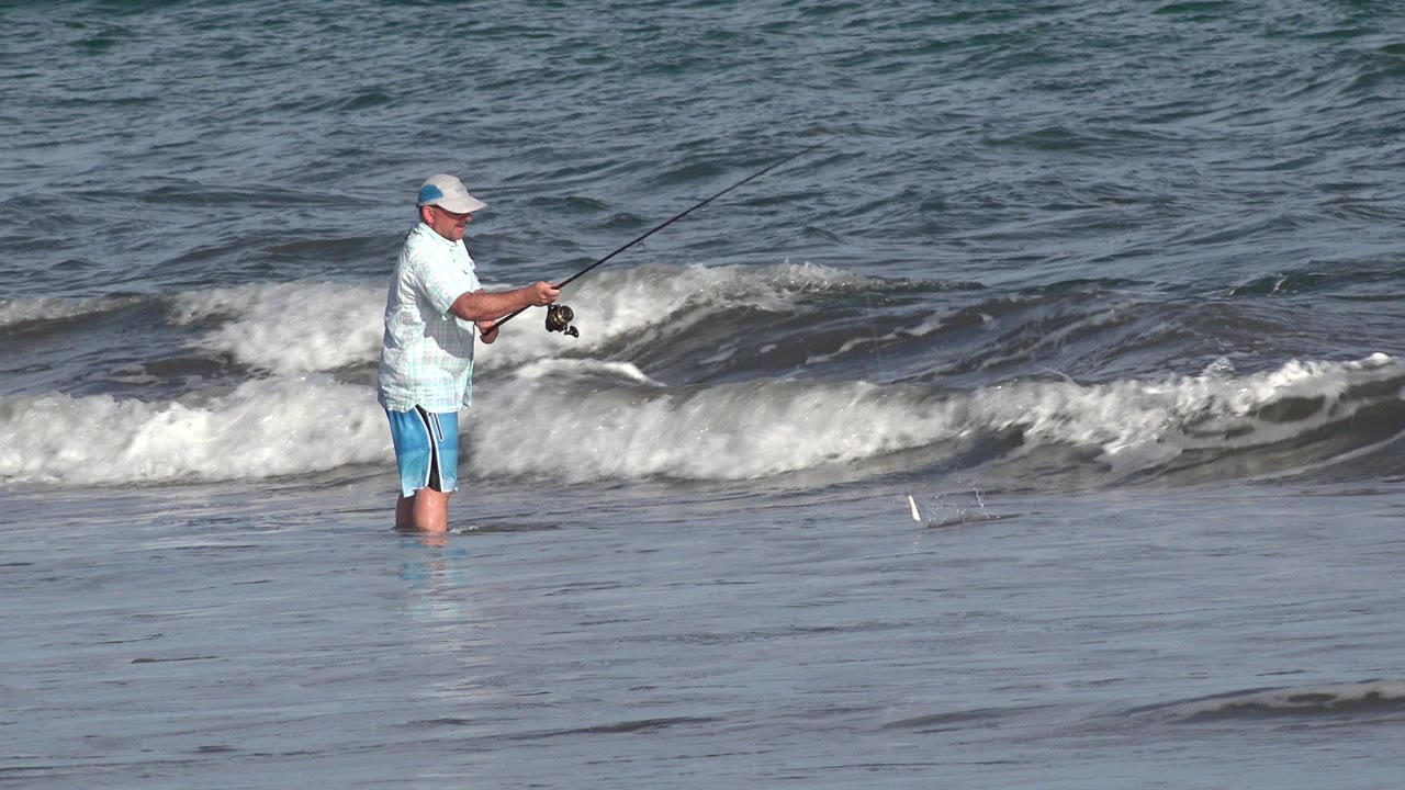 Adult man in a cap and light shirt fishes in shallow Los Cabos ocean water while a low wave folds across the background.